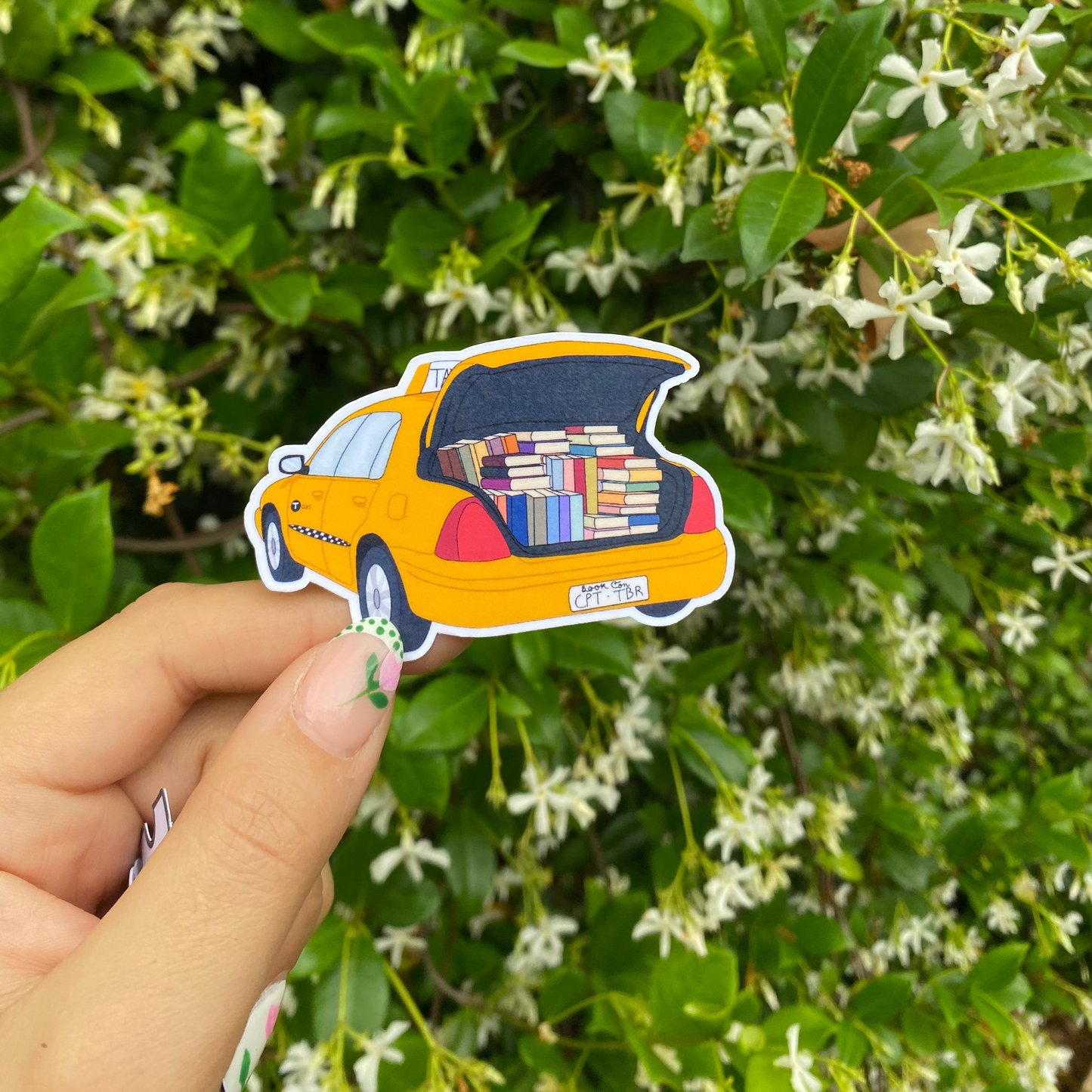 Hand holding a sticker of a yellow taxi with books open against a green leafy background